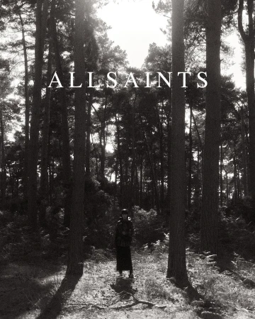 Monochrome photograph features a single female figure in a hat and coat standing in a dense pine forest in full-length shot.