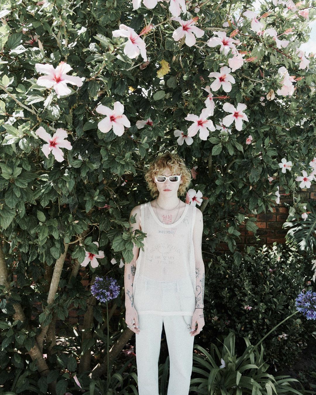 Young adult stands in front of vibrant pink hibiscus blossoms, wearing white sunglasses, top, and pants in an outdoor garden setting.
