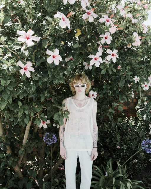 Young adult stands in front of vibrant pink hibiscus blossoms, wearing white sunglasses, top, and pants in an outdoor garden setting.