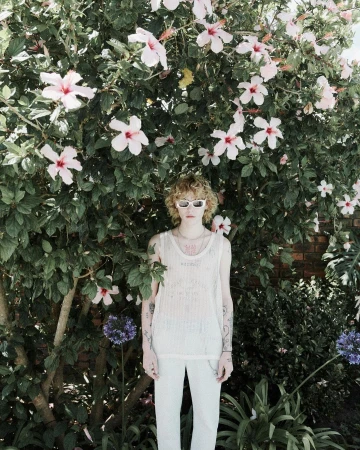Young adult stands in front of vibrant pink hibiscus blossoms, wearing white sunglasses, top, and pants in an outdoor garden setting.