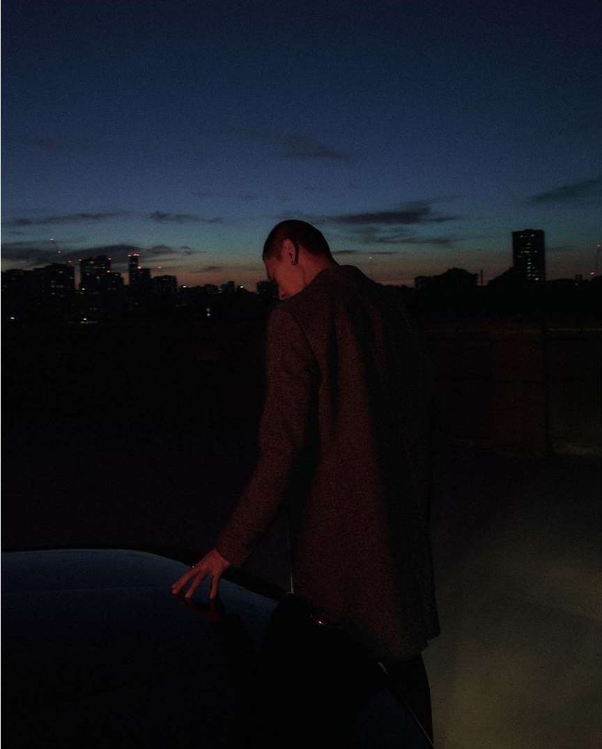 Man standing on a rooftop at dusk touching the hood of a car with a city skyline visible in the distance