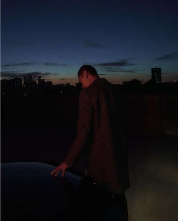 Man standing on a rooftop at dusk touching the hood of a car with a city skyline visible in the distance
