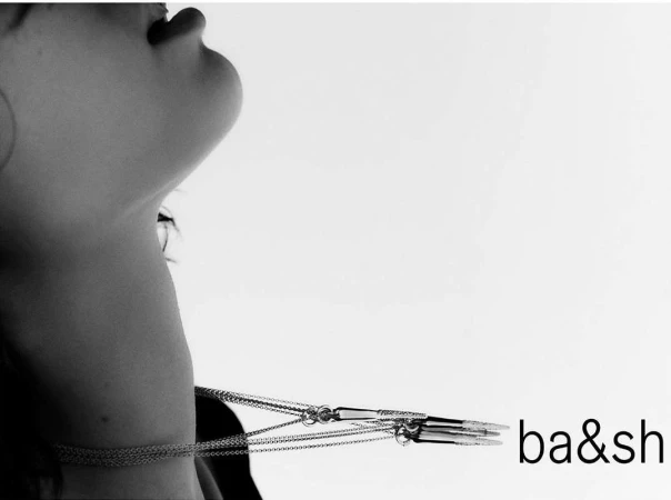 Fashion shot: An upward view showcases a fair-skinned person with layered necklaces, captured in high-contrast black and white, with the 'ba&sh' logo.