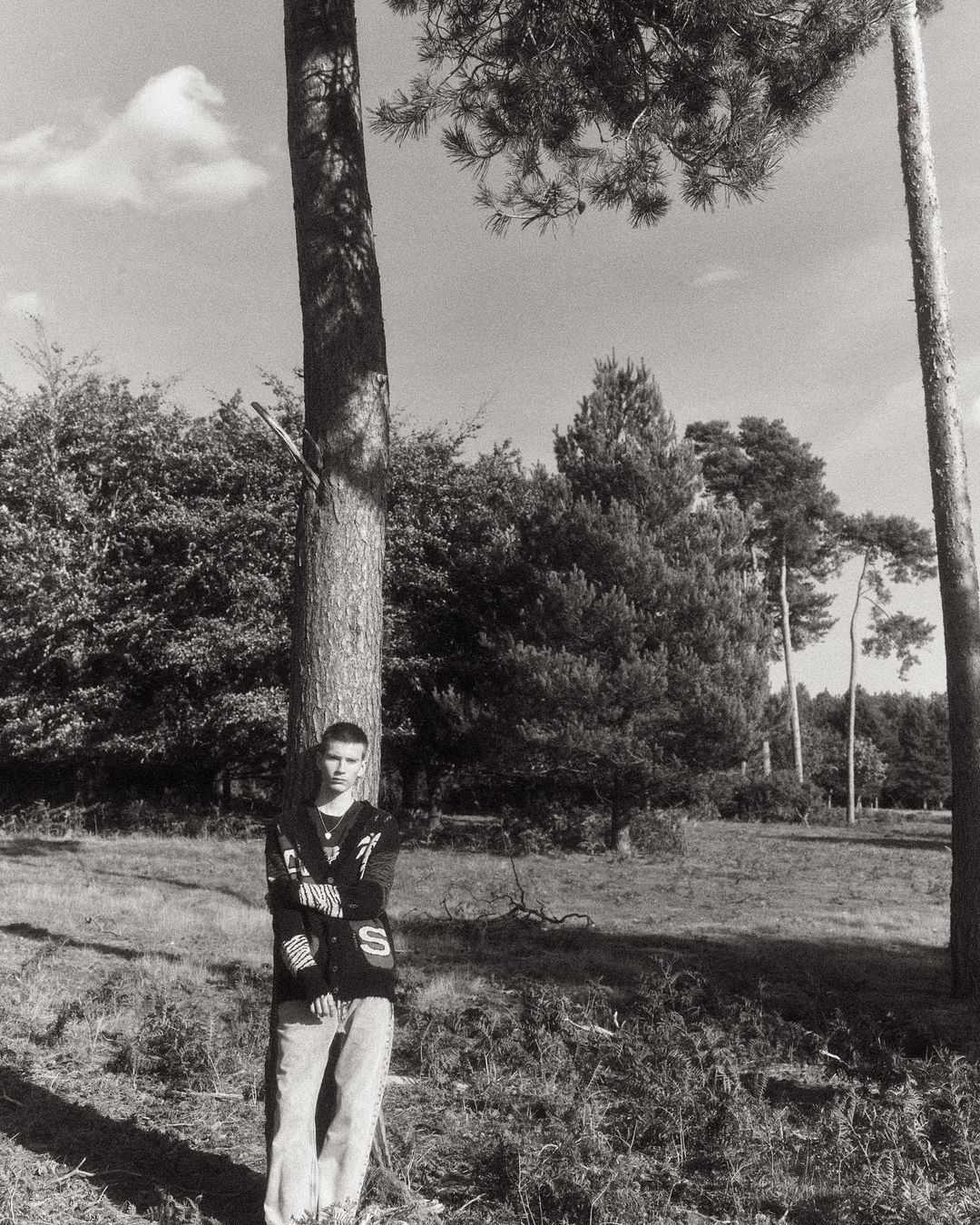 A young man leaning against a tall pine tree in a wooded landscape, photographed in monochrome.