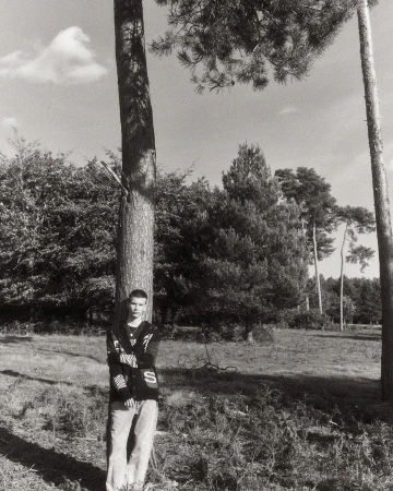 A young man leaning against a tall pine tree in a wooded landscape, photographed in monochrome.