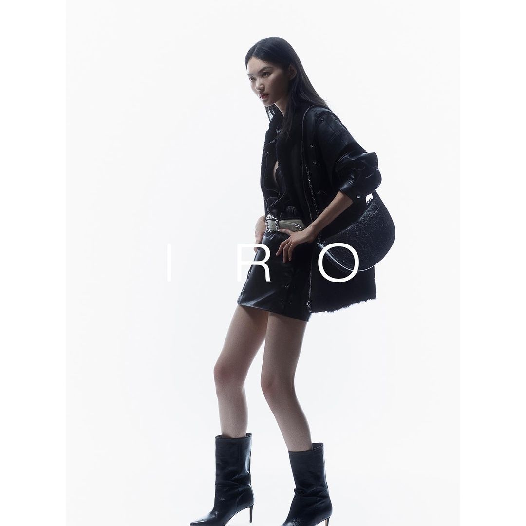 Model in black leather jacket and miniskirt holding a black handbag against a bright white seamless background