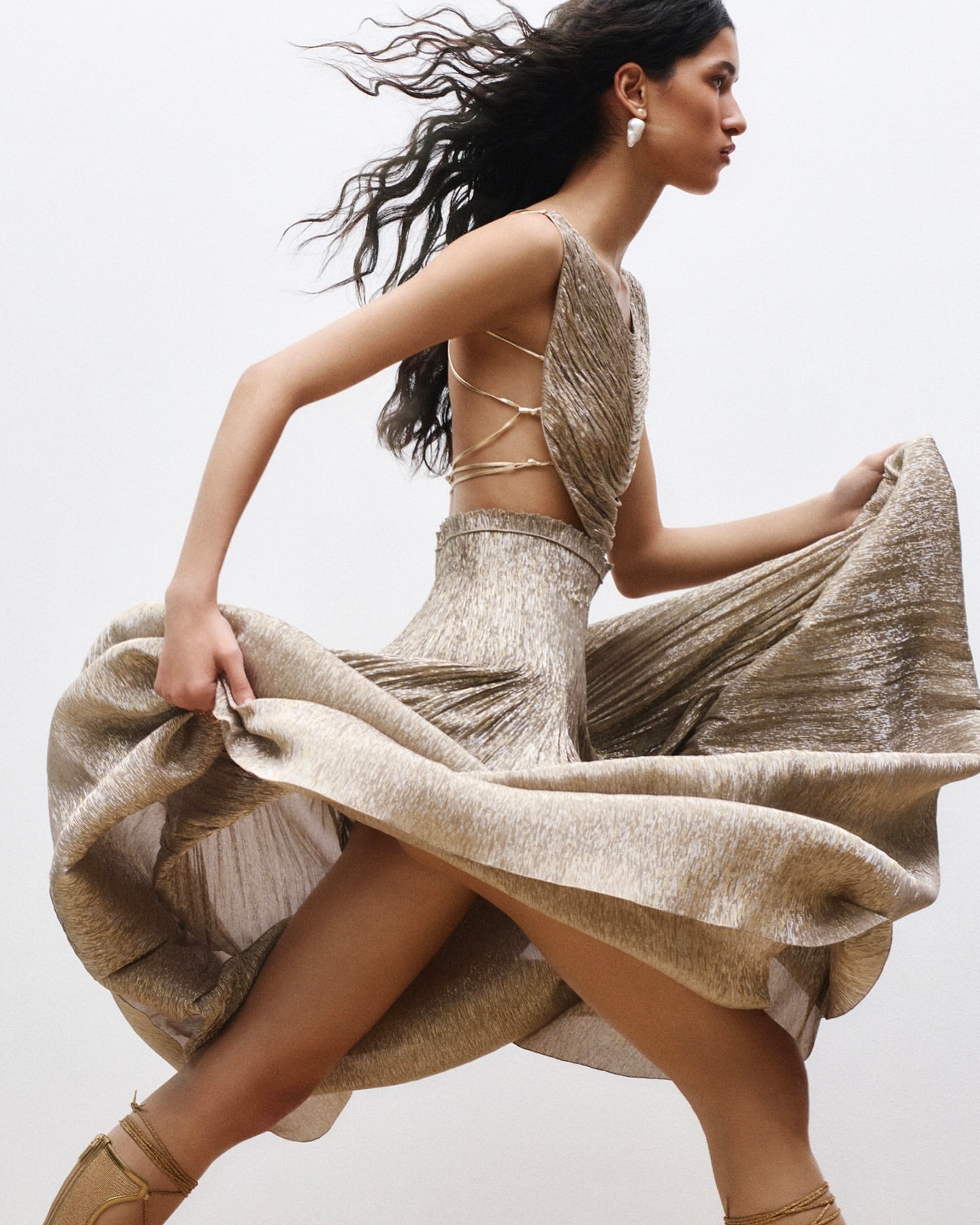 Fashionable woman in motion wearing a gold pleated open back dress and strappy heels against a plain background in a studio shot.