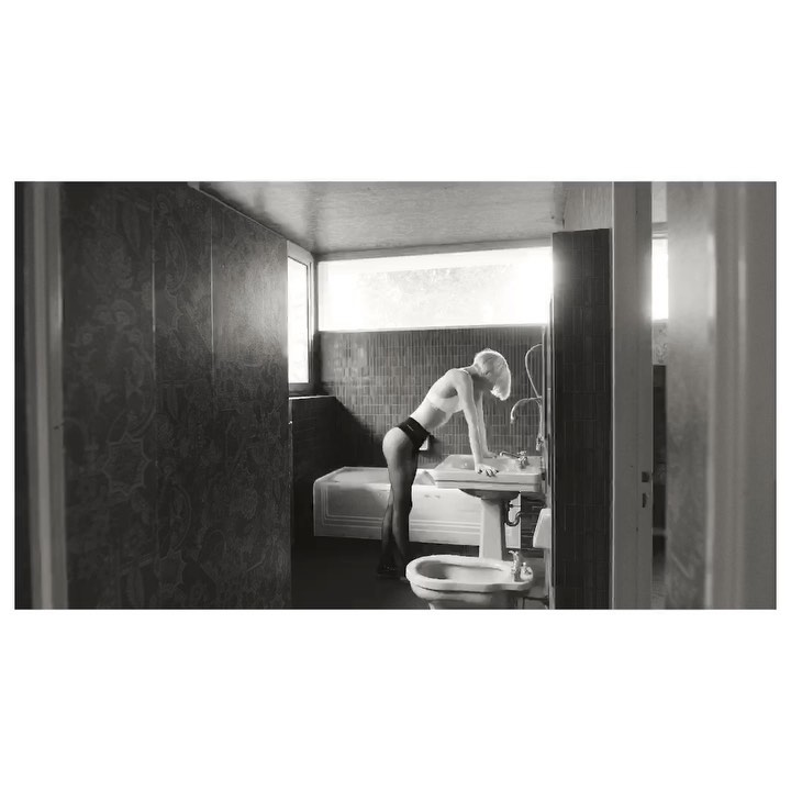 Monochrome photo of a slender, fair-skinned woman in minimal clothing leaning towards a sink in a tiled bathroom with natural light.