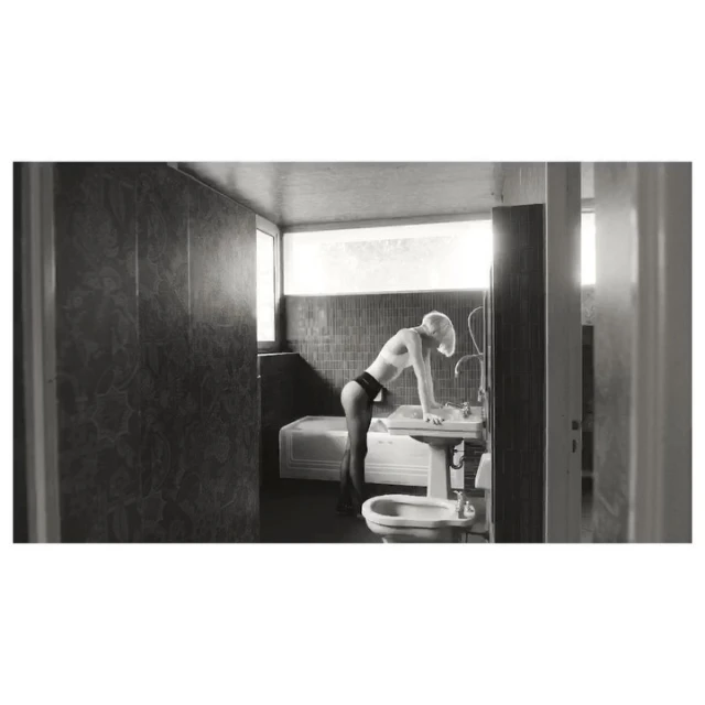 Monochrome photo of a slender, fair-skinned woman in minimal clothing leaning towards a sink in a tiled bathroom with natural light.