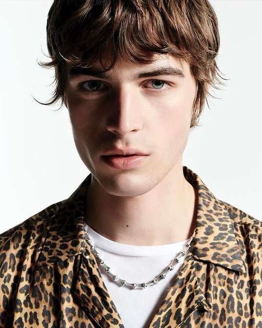Close-up portrait of a young man with brown hair wearing a leopard print shirt and silver chain necklace against a white background.