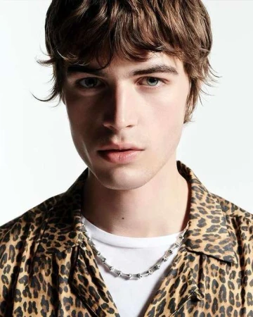 Close-up portrait of a young man with brown hair wearing a leopard print shirt and silver chain necklace against a white background.