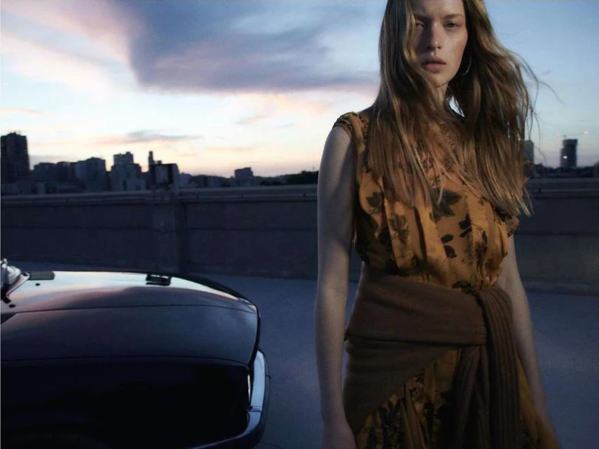 Woman poses in floral dress next to a black SUV at dusk with city skyline in background. Fashion editorial outdoor shot.