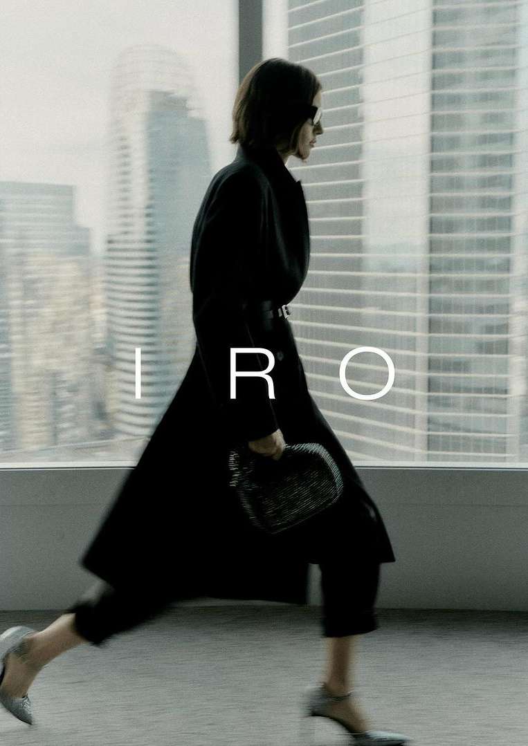 Fashion shot of a woman in a black coat, pants, and heels walking past a window in a city, holding a purse; IRO logo visible.