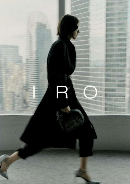 Fashion shot of a woman in a black coat, pants, and heels walking past a window in a city, holding a purse; IRO logo visible.