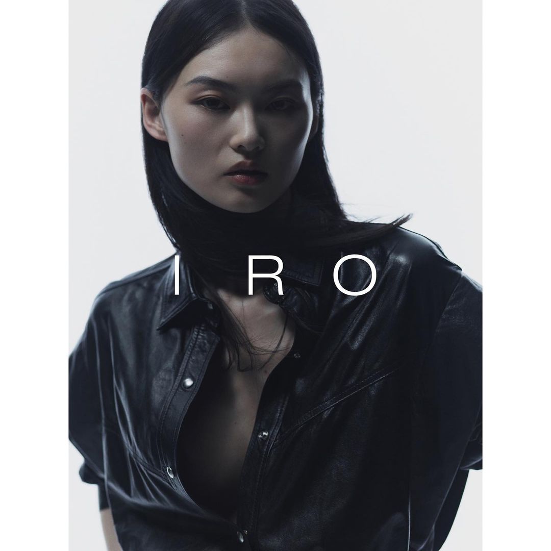 Fashion portrait of an Asian woman with dark hair, wearing a black leather-look shirt, against a bright white background with "IRO" text.