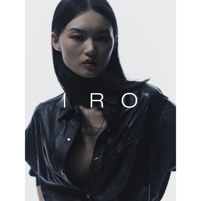 Fashion portrait of an Asian woman with dark hair, wearing a black leather-look shirt, against a bright white background with "IRO" text.