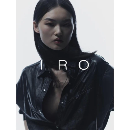 Fashion portrait of an Asian woman with dark hair, wearing a black leather-look shirt, against a bright white background with "IRO" text.
