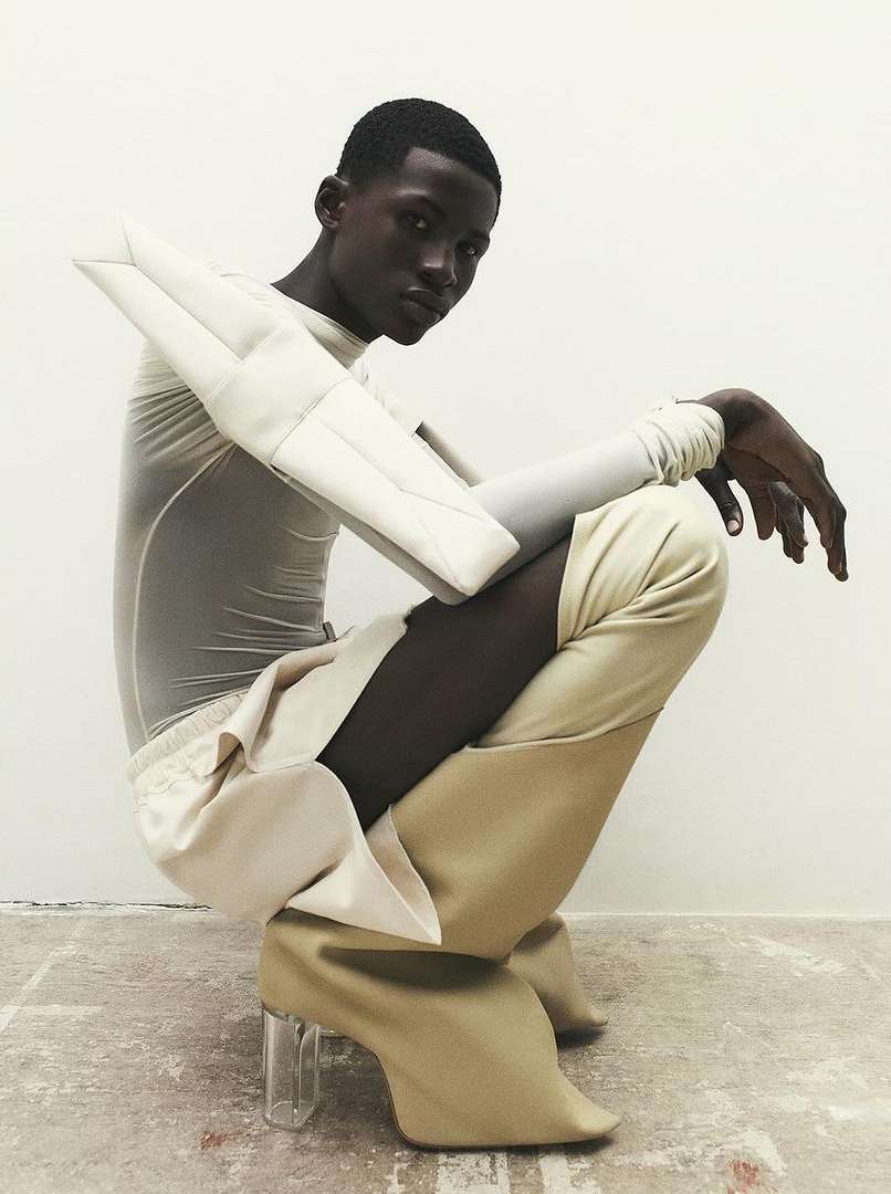A person with dark skin poses crouching in high-heeled boots, a long sleeve top and arm coverings against a light gray background.