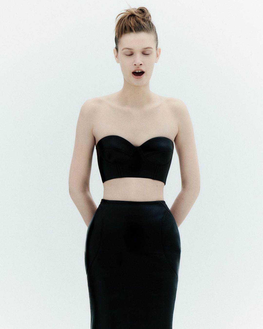 Elegant adult woman in a black two-piece outfit with her eyes closed in a bright indoor setting, capturing minimalist fashion.