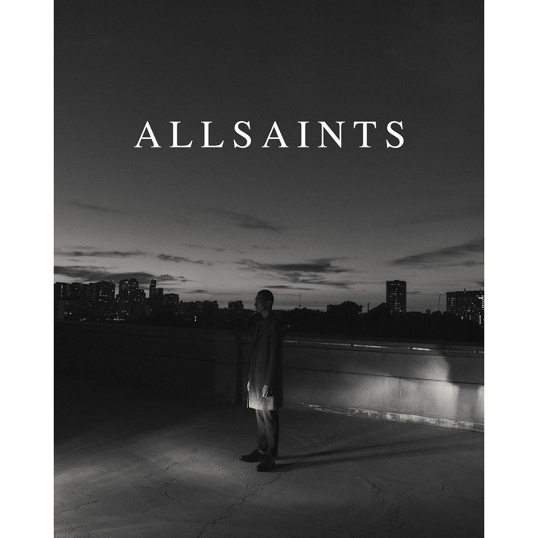 Monochrome fashion shot: a man in a dark coat stands on a rooftop at twilight with a city skyline, illuminated by a spotlight.