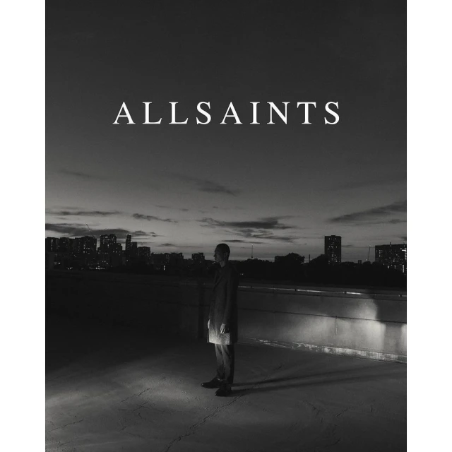 Monochrome fashion shot: a man in a dark coat stands on a rooftop at twilight with a city skyline, illuminated by a spotlight.