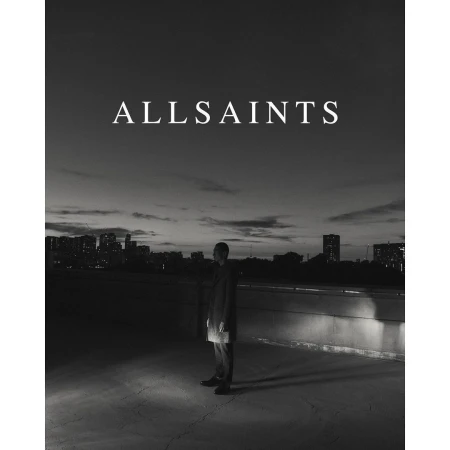 Monochrome fashion shot: a man in a dark coat stands on a rooftop at twilight with a city skyline, illuminated by a spotlight.