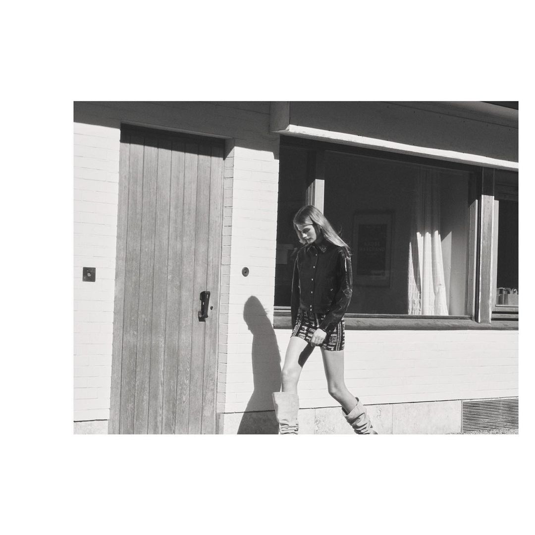 Monochrome portrait of a woman in a short plaid skirt, button-down shirt, and high socks posing next to a building with a door and window.