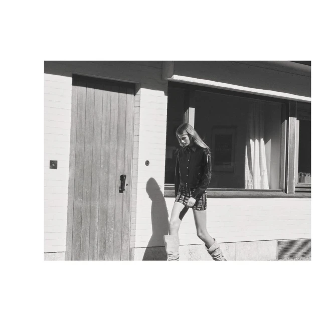 Monochrome portrait of a woman in a short plaid skirt, button-down shirt, and high socks posing next to a building with a door and window.