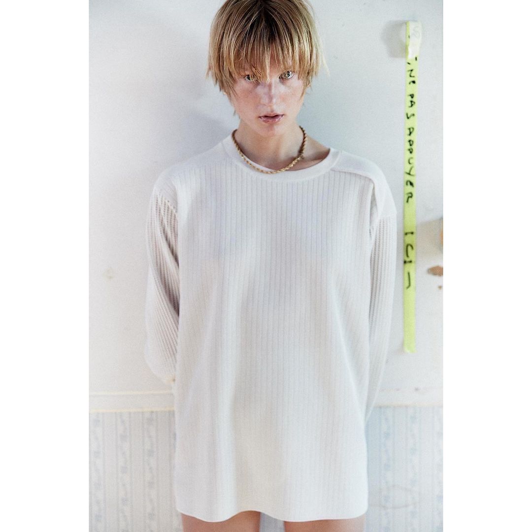 A blond-haired person wears a long-sleeved, vertically-ribbed white top and a necklace while standing indoors against a peeling wall.