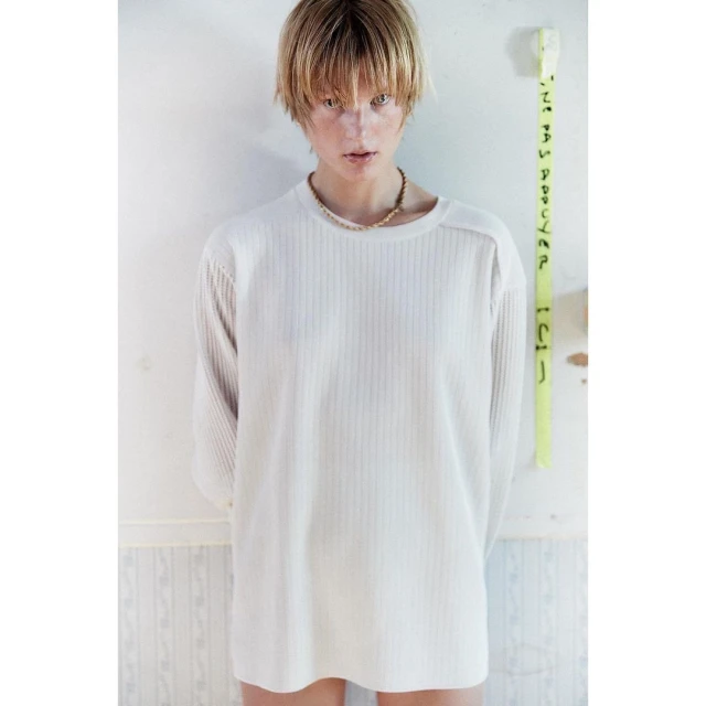 A blond-haired person wears a long-sleeved, vertically-ribbed white top and a necklace while standing indoors against a peeling wall.