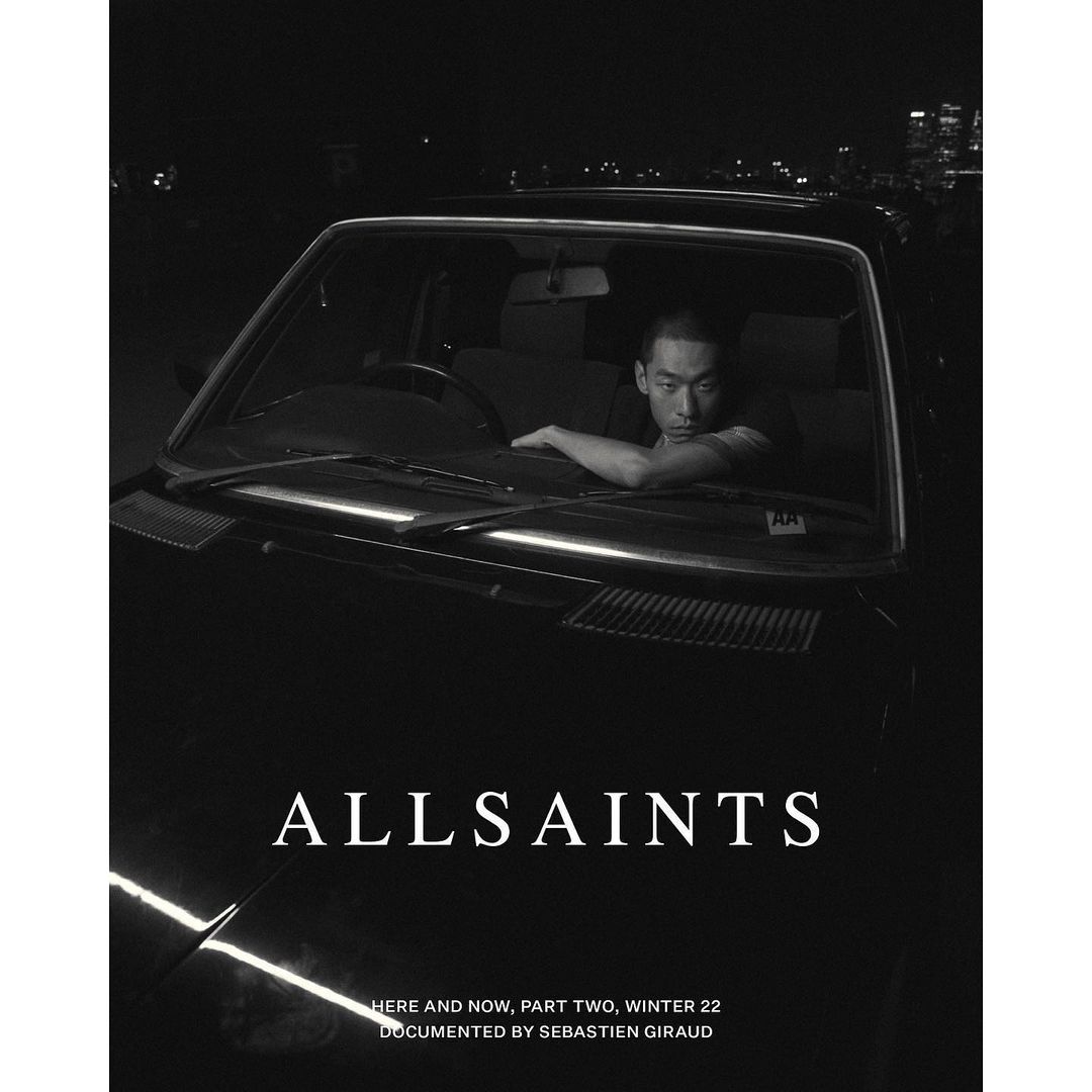 Black and white fashion-editorial shot of an adult male in a car, against an urban cityscape at night with visible brand logo.