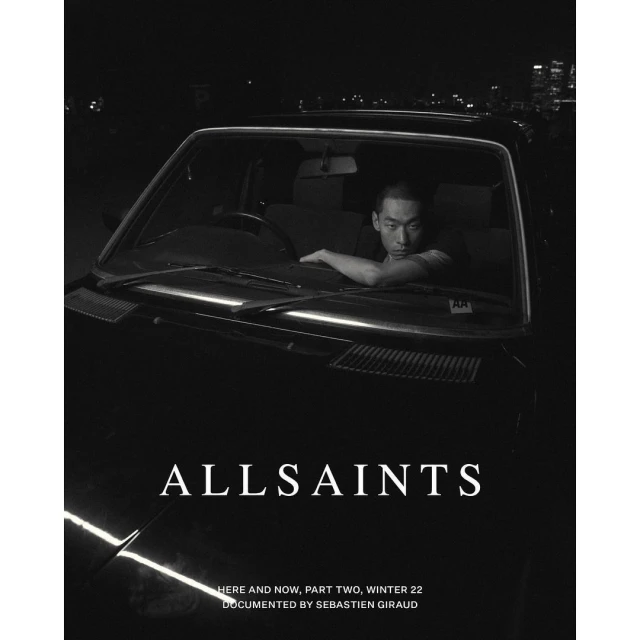 Black and white fashion-editorial shot of an adult male in a car, against an urban cityscape at night with visible brand logo.