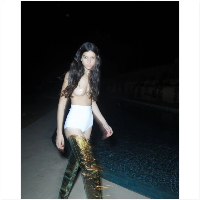 A woman with long hair wearing high-waisted white shorts and tall gold boots poses next to a dark pool at night.