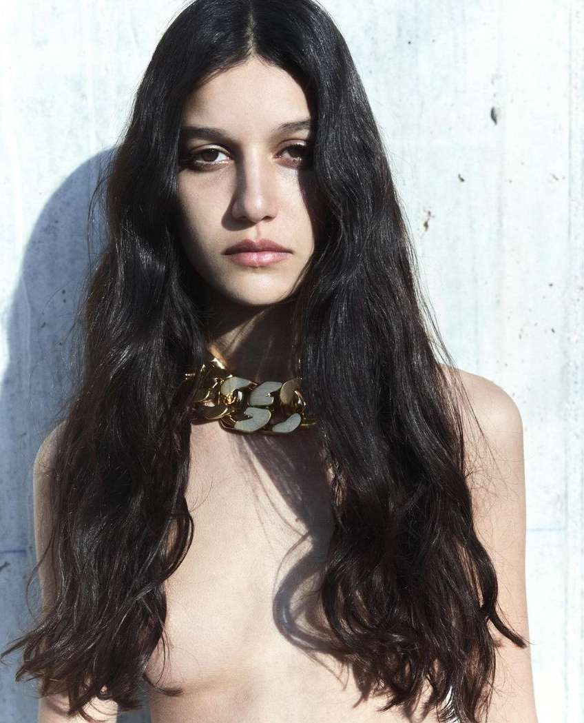 Portrait of a young adult woman with long dark hair wearing a chunky gold and white necklace, posing against a concrete wall.