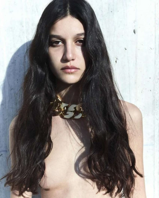 Portrait of a young adult woman with long dark hair wearing a chunky gold and white necklace, posing against a concrete wall.