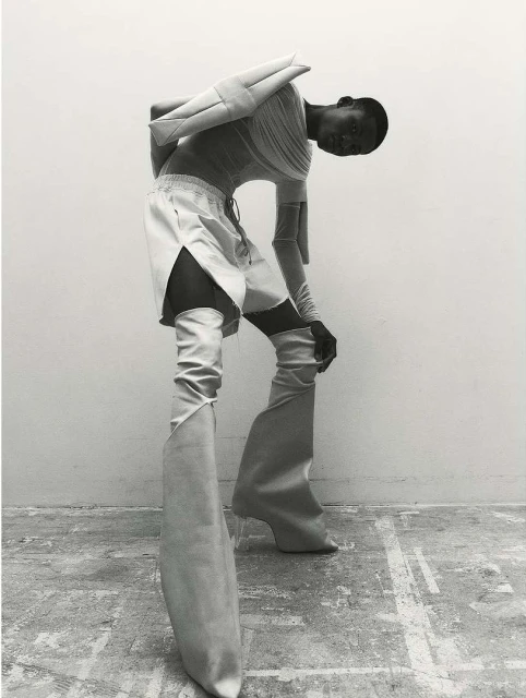 A person in avant-garde fashion, including sculptural shoulder piece, tunic, shorts, and high boots with clear heels, poses indoors.