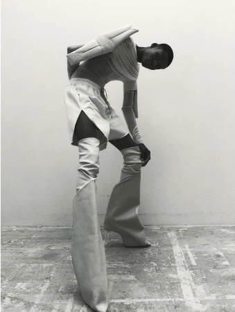 A person in avant-garde fashion, including sculptural shoulder piece, tunic, shorts, and high boots with clear heels, poses indoors.
