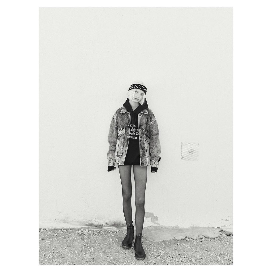 Monochrome shot of a blonde adult woman in an acid-wash jean jacket, black hooded sweatshirt, black tights, and boots, posing against a wall.