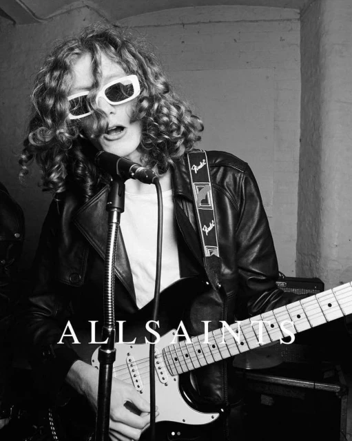 Black and white studio shot of a singer with curly hair wearing sunglasses and a leather jacket, playing an electric guitar and singing.