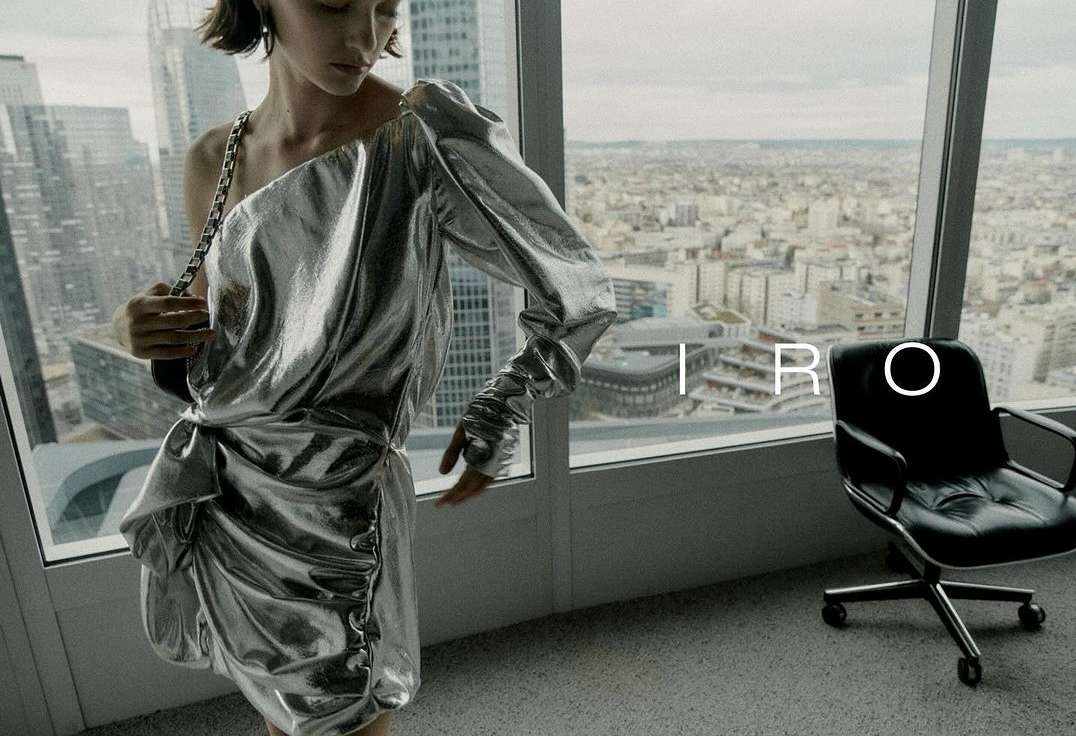 Elegant woman poses indoors wearing a silver, asymmetrical dress against a backdrop of a city skyline visible through large windows.