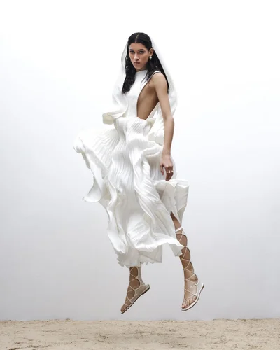 Model in white pleated dress with hood and lace-up sandals jumping against a neutral background