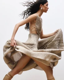 Model in a metallic gold pleated dress with long dark hair blowing in motion against a white backdrop.