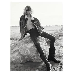 Model with long hair wearing an open studded denim jacket and cowboy boots seated on a rock outdoors.