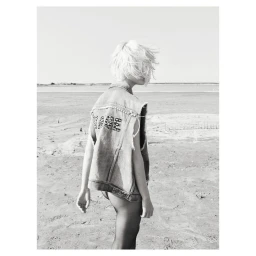 Monochrome fashion portrait of a short-haired blonde woman wearing an embroidered denim vest outdoors on a sandy beach landscape.