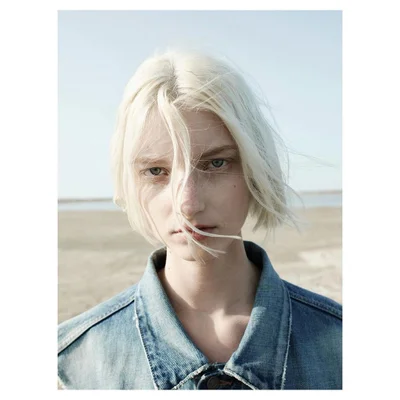 Portrait of a young model with short platinum blonde hair and a denim jacket against an outdoor sandy background.