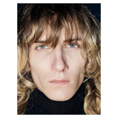 Close-up portrait of a man with long blonde curly hair and blue eyes wearing a black turtleneck against a dark background.