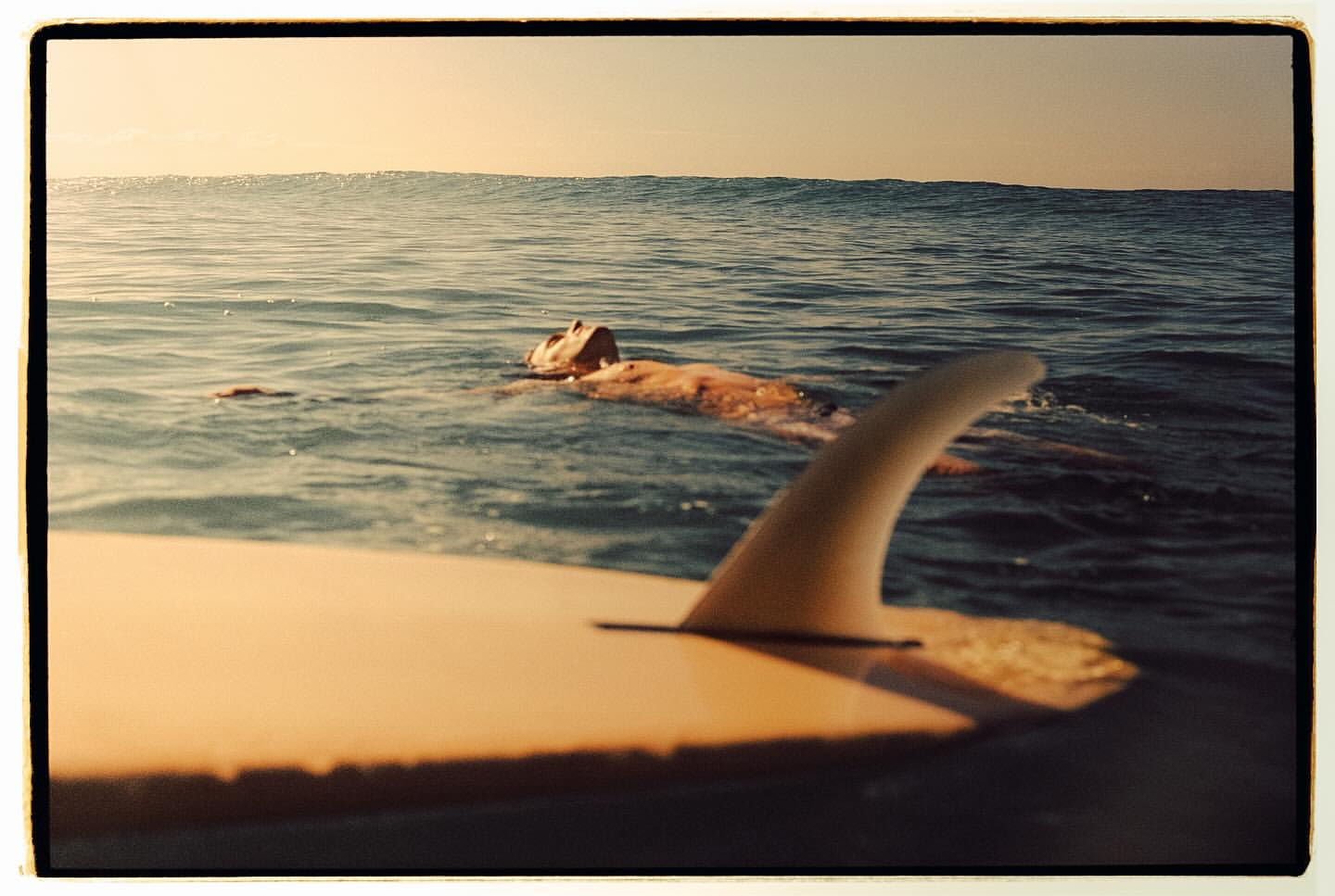 A surfer floats in calm ocean water with a surfboard in the foreground, under a clear sky, creating a serene summer scene.