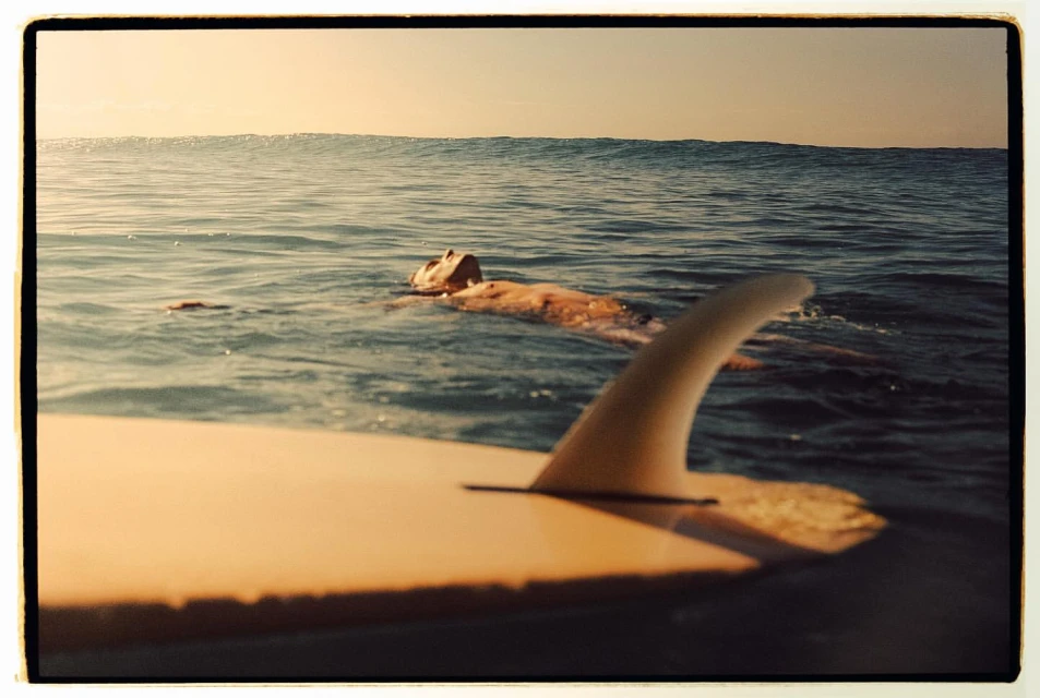 A surfer floats in calm ocean water with a surfboard in the foreground, under a clear sky, creating a serene summer scene.