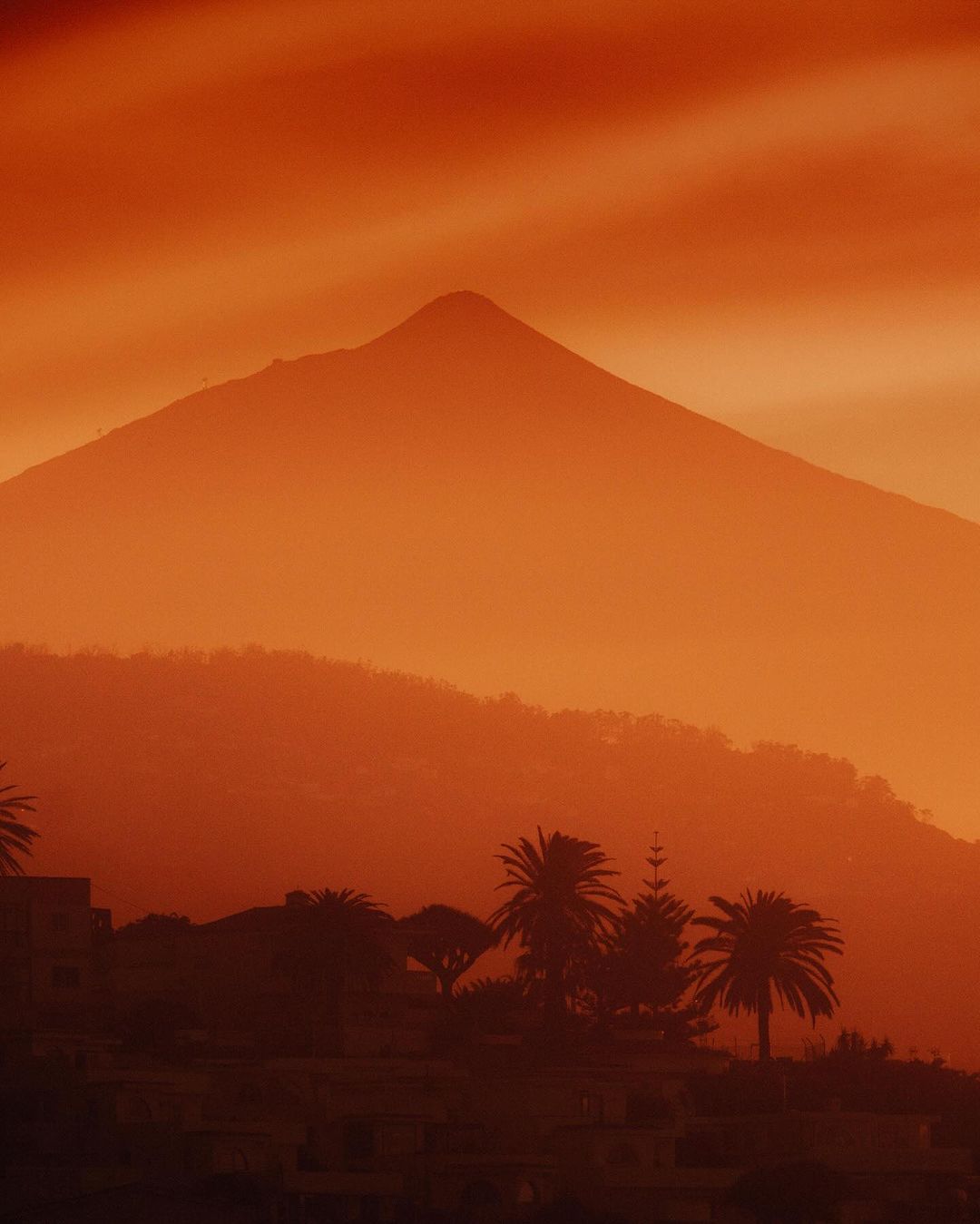 Orange-toned scenic shot of a mountain range and silhouette palm trees in the foreground, creating a warm and tropical landscape.