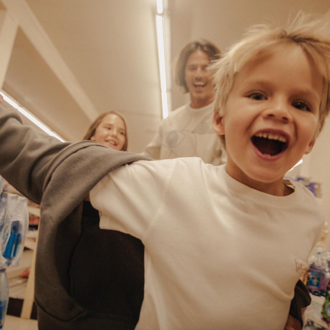 A cheerful young boy smiles broadly while being pushed in a shopping cart in a supermarket aisle, with two other people smiling behind him.